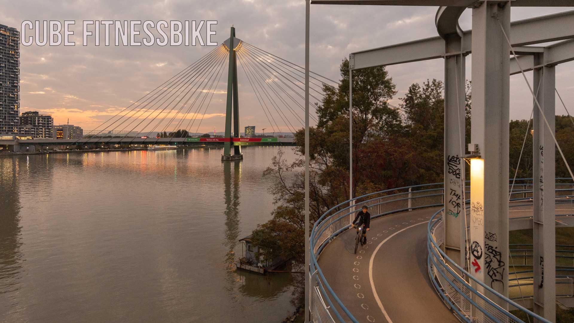 On voit une ville au bord d'un fleuve. Au premier plan, une cycliste roule sur la piste cyclable au crépuscule. L'image est intitulée Cube Fitnessbike.