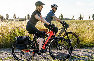 On voit un couple roulant à vélo électrique sur un chemin de gravier. En arrière-plan, on aperçoit de hautes herbes.