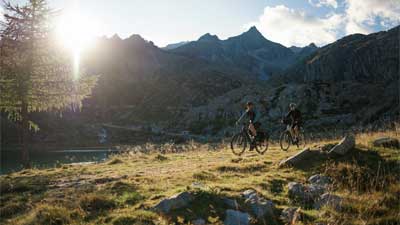 Un couple roule à vélo sur un sentier de randonnée. En arrière-plan, on aperçoit des montagnes derrière lesquelles le soleil se lève.