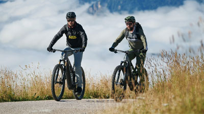 Deux cyclistes roulent sur leur vélo électrique tout terrain sur une piste cyclable. Des montagnes sont visibles en arrière-plan.
