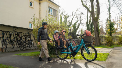 On voit une mère qui court vers son vélo cargo bleu. Deux petits enfants sont assis sur le porte-bagages. La scène se déroule dans un quartier résidentiel.