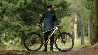 On voit un homme derrière son VTT Cube doré, sur un chemin forestier.