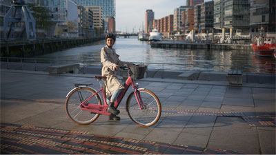 On voit une femme sur un vélo électrique rose Cube Ella Hybrid. Elle roule le long d'une rivière. Une ville est visible en arrière-plan.