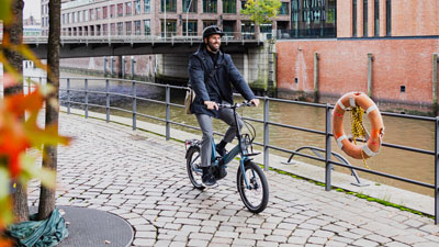 Un homme roule sur son vélo pliable bleu, avec des maisons et une rivière en arrière-plan.
