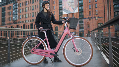 On voit une femme derrière un vélo électrique rose Cube Ella Hybrid. Un bâtiment apparaît à l'arrière-plan.