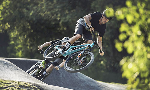 Un cycliste saute avec un dirt bike bleu clair dans un bike park, un autre cycliste avec un vélo vert à l'arrière-plan.