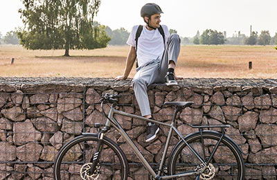 On voit un homme casqué, assis sur un mur. Au premier plan, il y a un vélo gris et, à l'arrière-plan, un paysage champêtre.