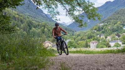 Un cycliste roule sur un chemin de gravier avec son VTT, avec un paysage montagneux en arrière-plan.
