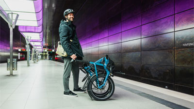 On voit un homme dans une station de métro, devant lui se trouve un vélo pliant bleu replié.