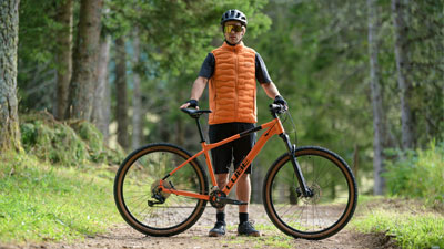 Un homme se tient derrière son VTT orange Cube, sur un chemin forestier.