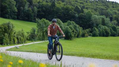 Un cycliste roule sur une piste cyclable goudronnée avec son VTT, avec un paysage rural en arrière-plan.