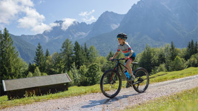 Un garçon roule à VTT sur un chemin caillouteux à travers un paysage montagneux. Une cabane est visible à l'arrière-plan. 