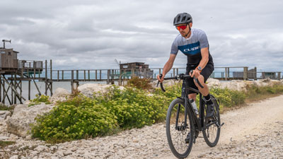 On voit un homme en tenue de cycliste. Il roule sur son vélo de gravel sur une piste cyclable le long d'une plage.