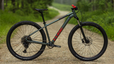 On voit une femme vêtue d'une tenue de cycliste violette, devant elle se trouve un VTT violet. En arrière-plan, on aperçoit un paysage montagneux.