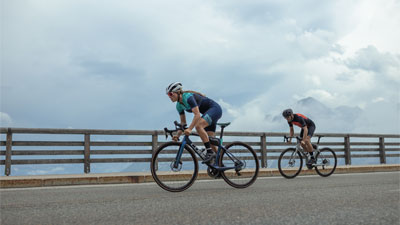 On voit deux cyclistes sur des vélos de course, avec une clôture en bois et des nuages en arrière-plan.
